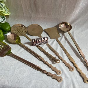 May include: A collection of copper-colored kitchen utensils, including a slotted spoon, ladle, and spatula, arranged on a white surface. The handles have a textured design. The image also includes green bell peppers and a tomato.