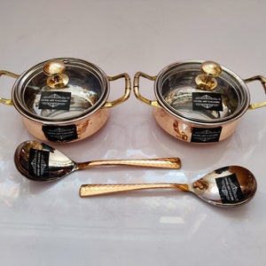 May include: Two small copper serving bowls with lids and two matching copper spoons. The bowls have a hammered finish and gold handles. The lids are made of clear glass.
