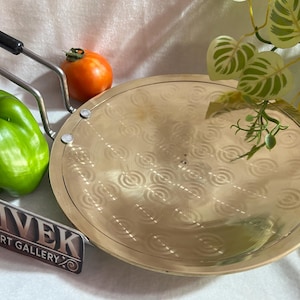 Puede incluir: Una plancha redonda dorada con un diseño decorativo, acompañada de un pimiento verde, un pequeño tomate naranja y un par de pinzas con mango negro. La imagen también incluye un letrero que dice "VIVEK ART GALLERY".