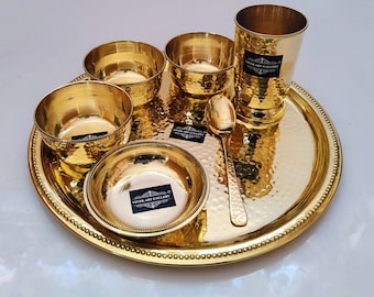 May include: A set of gold-toned brass dinnerware with a hammered finish. The set includes a large plate, four bowls, a glass, and a spoon. The set is arranged on a white surface.