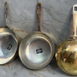 May include: Three hammered metal frying pans with long handles. The pans are silver, gold, and a combination of silver and gold.
