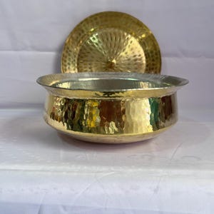 Puede incluir: Un cuenco de latón martillado con una tapa a juego. El cuenco y la tapa tienen un acabado dorado brillante y una superficie texturizada.