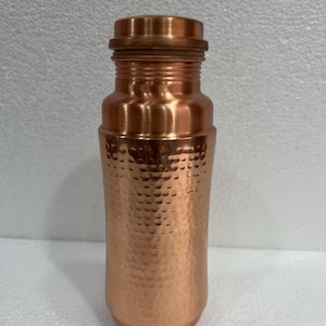 May include: A copper-coloured water bottle with a hammered texture on the body. The bottle has a screw-on lid and a rounded base. The overall design is simple and elegant, suitable for everyday use. The bottle is approximately 25 cm tall.