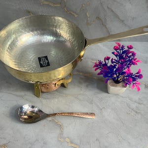 Puede incluir: Un bol de servir de latón martillado con un mango largo, sentado sobre un soporte de cobre. El bol está sobre una superficie de mármol blanco con una pequeña disposición de flores rosas y moradas en una maceta blanca.