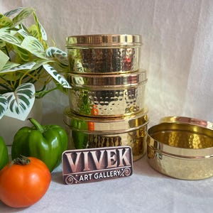 May include: Four gold-coloured, hammered-metal containers with lids are stacked, with an open container beside them. A sign reading "VIVEK ART GALLERY" is in front. Green peppers, a tomato, and a plant are also in the image.