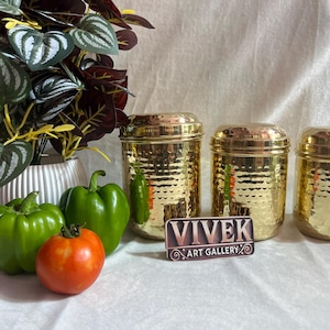 Puede incluir: Tres botes dorados con textura martillada y tapas. Pimientos verdes y un tomate rojo están frente a los botes. Una planta en maceta con hojas verdes y burdeos está en el fondo. El letrero dice "VIVEK ART GALLERY".