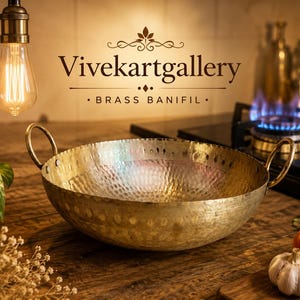 May include: A brass pan with a hammered finish and two handles is on a wooden surface. A lit gas hob and a chopping board with a tomato and garlic are in the background. The text "Vivekartgallery" and "Brass Banifil" are visible.