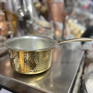 Puede incluir: Una pequeña cacerola dorada martillada con un mango largo. La olla tiene un interior plateado y se encuentra sobre una superficie metálica. El fondo está borroso, mostrando otros utensilios de cocina.