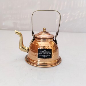 Puede incluir: Una pequeña tetera de cobre con un acabado martillado y un pico y un asa de latón. La tetera tiene una tapa y una pegatina que dice "Vivek Art Gallery".