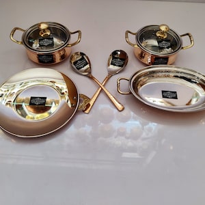 May include: A set of copper and stainless steel serving dishes with glass lids. The set includes two small bowls, a large oval dish, a round dish, and two serving spoons.
