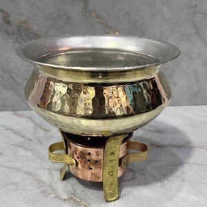 Puede incluir: Un calentador de alimentos de cobre y latón martillado con un cuenco redondo y un soporte de tres patas. El cuenco tiene un interior plateado.