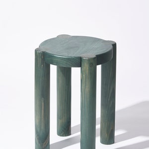 May include: A teal-colored wooden stool with a round seat and four cylindrical legs. The legs are evenly spaced and attached to the seat with rounded corners. The wood grain is visible, adding texture to the design. The stool is a simple, modern piece of furniture.