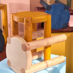 K&ouml;nnte beinhalten: Ein Holzhocker mit einer hellbraunen Oberseite und gelb gebeizten Beinen. Der Hocker ist aus Naturholz gefertigt und hat eine abgerundete Form. Der Hocker steht auf einer blauen Oberfl&auml;che.