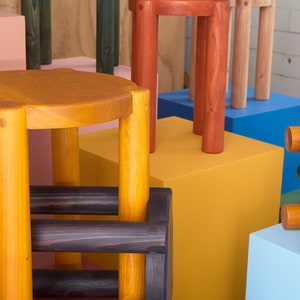K&ouml;nnte beinhalten: Eine Sammlung von Holzhockern in verschiedenen Farben, darunter Gelb, Orange, Gr&uuml;n und Blau. Die Hocker sind aus Naturholz gefertigt und haben ein schlichtes, minimalistisches Design.