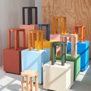 K&ouml;nnte beinhalten: Eine Sammlung von Holzhockern in verschiedenen Farben, darunter Rot, Blau, Gr&uuml;n, Gelb und Naturholz. Die Hocker sind auf bunten Bl&ouml;cken angeordnet und bilden ein optisch ansprechendes Display.