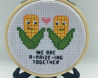 May include: A white embroidery hoop with a cross-stitch design of two corn cobs with faces, holding hands with a pink heart between them. The text below reads "We are a-maize-ing together".