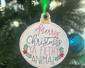 May include: A white Christmas ornament with green ribbon. The ornament is embroidered with the text "Merry Christmas Ya Filthy Animal" in red and black thread. The ornament is decorated with green holly leaves and red berries.