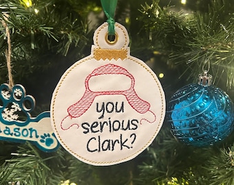 May include: A white Christmas ornament with red and gold embroidery. The ornament features a red hat with ear flaps and the text "you serious Clark?"