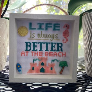 May include: White framed cross-stitch with the text "Life is always better at the beach". The cross-stitch features a yellow sun, a blue and green surfboard, a pink sandcastle, a green palm tree, and a blue seahorse.