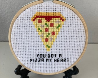 May include: A white embroidery hoop with a cross-stitch design of a pizza slice in the shape of a heart. The text "You got a pizza my heart" is stitched below the pizza slice.