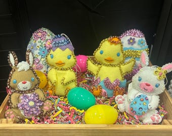 May include: A wooden crate filled with colorful Easter decorations, including two yellow chicks, two bunnies, and several decorated eggs. The decorations are made of felt and covered in sequins.