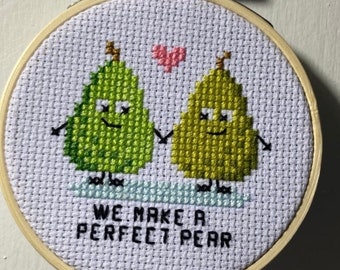 May include: A white embroidery hoop with a cross-stitch design of two green pears holding hands with a pink heart above them. The text "We make a perfect pear" is stitched below the pears.