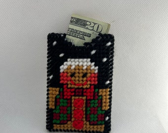 May include: A handmade, black, cross-stitched wallet featuring a gingerbread man design. The gingerbread man has a tan body, red scarf, and green and red plaid jacket. A twenty-dollar bill is visible inside the wallet.