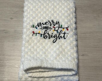 May include: White kitchen towel with the text "merry and bright" embroidered in black with a string of colorful lights above the text.