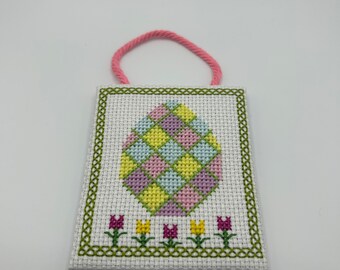 May include: A white cross-stitch ornament with a pink, yellow, and blue egg design and a row of pink and yellow tulips at the bottom. The ornament has a pink loop for hanging.