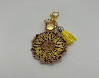 May include: A yellow sunflower embroidered on a brown leather keychain with a gold metal clasp and a yellow tassel.
