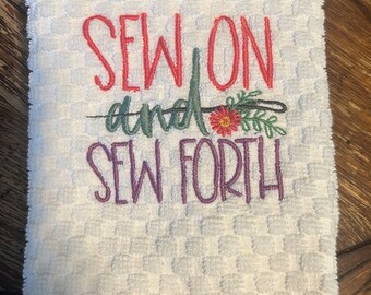 May include: White kitchen towel with embroidered text "Sew on and sew forth" in red and purple thread. The embroidery includes a small floral design with a needle and thread.