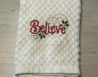 May include: White kitchen towel with a red embroidered design that says "Believe" with green leaves and gold swirls.