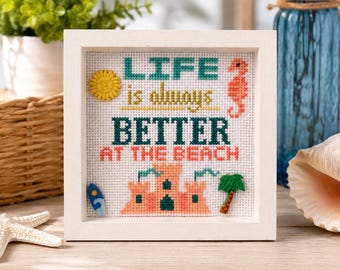 May include: A square white wooden frame containing a cross-stitch piece with the text "LIFE is always BETTER AT THE BEACH". The design includes a yellow sun, an orange seahorse, a blue surfboard, a peach sandcastle, and a green palm tree.