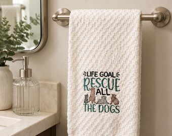 May include: A white waffle-weave hand towel featuring embroidered text that reads LIFE GOAL RESCUE ALL THE DOGS in teal and black thread, accompanied by small embroidered illustrations of four dogs in various shades of brown and gray.