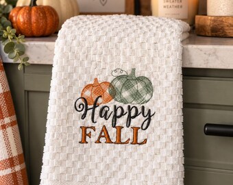May include: A white waffle-weave kitchen towel featuring an embroidered design of one orange plaid pumpkin and one green plaid pumpkin. Below the pumpkins, the text reads Happy FALL in black and orange script and block lettering respectively.