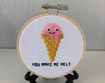 May include: A small, round, wooden embroidery hoop with a white fabric background. The fabric has a pink and yellow embroidered ice cream cone with a smiling face. The text "You make me melt" is embroidered below the ice cream cone.