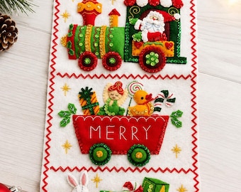 May include: A white felt wall hanging featuring a three-car train design with red zigzag stitching. The top car shows Santa in a green engine, the middle red car holds gifts and a duck with the word MERRY, and the bottom green car holds a rabbit and blocks with the word CHRISTMAS.