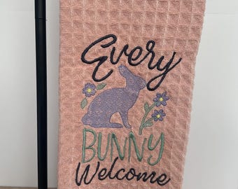 May include: A pink waffle weave kitchen towel with the embroidered text "Every Bunny Welcome" and a purple bunny silhouette with green and purple flowers.