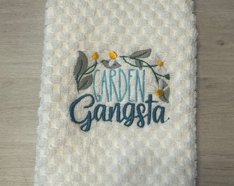 May include: White, textured hand towel with the embroidered text "Garden Gangsta" in teal script. The towel features a floral design with yellow and white accents, and green leaves.