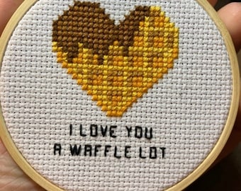 May include: A white embroidery hoop with a cross-stitch design of a heart-shaped waffle with the text "I love you a waffle lot".