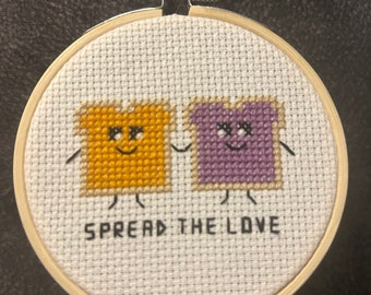 May include: A white embroidery hoop with a cross-stitch design of two cartoon bread slices holding hands. The bread slices are yellow and purple and have happy faces. The text "SPREAD THE LOVE" is stitched below the bread slices.