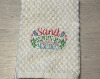 May include: White kitchen towel with embroidered text "Sand Between My Mistletoes" in pink, blue, and green.