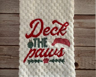 May include: White kitchen towel with red embroidered text that says "Deck the paws" with a green ornament, a red Santa hat, and a green paw print.