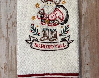 May include: White hand towel with an embroidered Santa Claus wearing a cowboy hat and boots. The design includes the text "HO HO HO Y'ALL" on a banner. The bottom of the towel has red and white stripes.