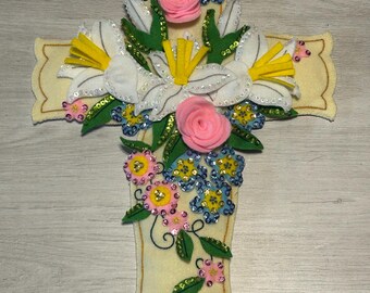 May include: A decorative, cream-colored felt cross adorned with fabric flowers and sequins. The cross features white lilies with yellow centers, pink roses, and clusters of blue and pink flowers. Green leaves and gold trim add detail.