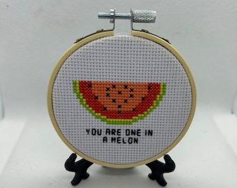 May include: A round wooden embroidery hoop with a white fabric background. The fabric has a cross-stitch design of a watermelon slice with the text "You are one in a melon". The hoop is on a black stand.
