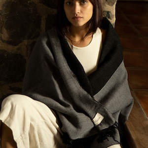 Może przedstawiać: Kobieta w białym topie na ramiączkach i białych spodniach siedzi przy kamiennej ścianie. Ma na sobie szal w szare i czarne pasy z frędzlami. Szal ma małą białą metkę z napisem "Fendi".