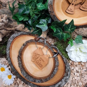 May include: Two wooden keychains with the text "Soll Bestet zum Gl&uuml;ck" and "Schl&uuml;ssel zum Gl&uuml;ck" on a wooden tray with a natural wood grain finish. The tray is surrounded by wood chips and green ivy.
