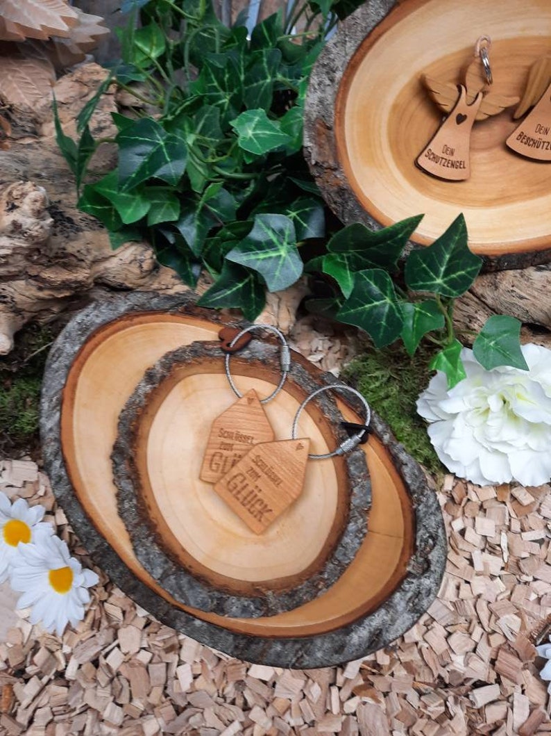 May include: Two wooden keychains with the text "Soll Bestet zum Gl&uuml;ck" and "Schl&uuml;ssel zum Gl&uuml;ck" on a wooden tray with a natural wood grain finish. The tray is surrounded by wood chips and green ivy.
