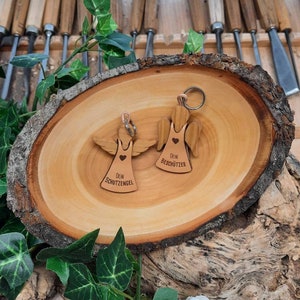 May include: Two wooden angel keychains with the text "Dein Schutzengel" and "Dein Besch&uuml;tzer" on them. The keychains are on a wooden surface.
